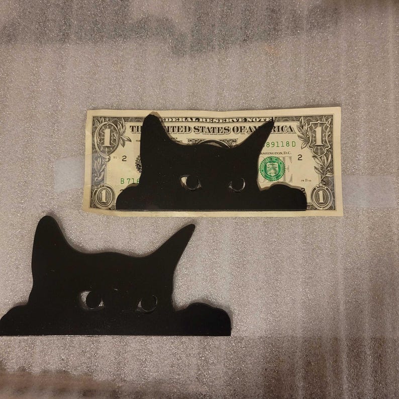 A photo with a smaller decorative cat placed on top of a dollar bill. The small cat is smaller than the dollar bill in width and height.