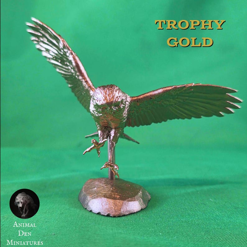 May include: A bronze-colored eagle figurine with outstretched wings, poised as if landing. The sculpture is mounted on a small, textured base. The text "TROPHY GOLD" is displayed above the eagle. The logo for "Animal Den Miniatures" is in the lower left corner.