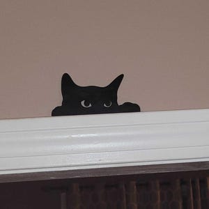 A 3D printed black cat decoration made of resin, designed to appear as if it is peeking over a door frame or shelf edge. The cat has large, detailed eyes and a sleek, smooth finish
