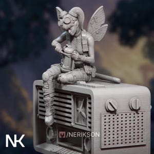 Op de afbeelding: Een grijze beeldje van een fee met een bril en vleugels, zittend op een vintage radio. De fee houdt een spuitbus vast. De radio heeft knoppen en een luidsprekerrooster. Het beeldje heeft ingewikkelde details.