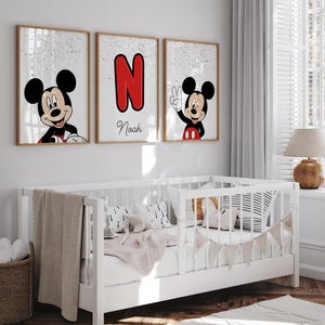 May include: Three framed Mickey Mouse art prints. One features Mickey smiling, another displays a red letter "N", and the third shows Mickey giving a peace sign. The name "Naah" is written below the letter "N". The prints are displayed above a white crib.