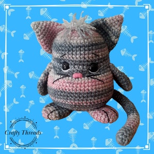 May include: A crocheted gray and pink cat toy with black eyes and a pink nose. The cat has a long tail and is sitting on a blue background with a fishbone pattern.