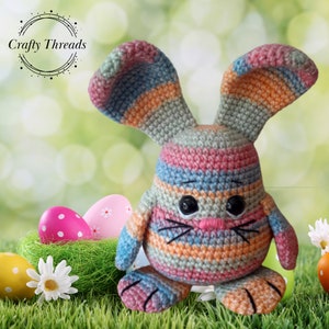 May include: A colorful crocheted Easter bunny with striped ears and a pink nose. The bunny is sitting in a nest of green grass with Easter eggs.