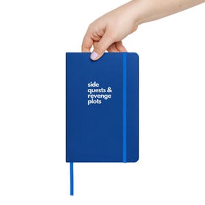May include: A blue notebook with a blue ribbon bookmark. The front cover has the text "side quests & revenge plots" in white.