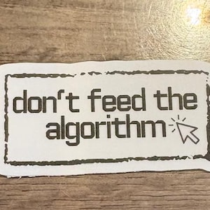 May include: A white rectangular sticker with a black border and the words "don't feed the algorithm" in a bold, sans-serif font. A computer cursor graphic is to the right of the text. The sticker is on a wooden surface.