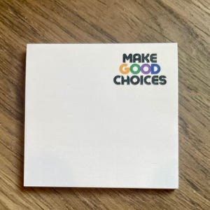 Könnte beinhalten: Ein weißer Haftnotiz mit dem Text "MAKE GOOD CHOICES" in schwarzen Buchstaben mit einem Regenbogen-Farbschema.