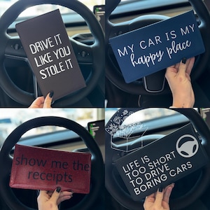 May include: Four different car dashboard organizers with funny sayings. The first is black with white text that says "Drive it like you stole it". The second is blue with white text that says "My car is my happy place". The third is red with white text that says "Show me the receipts". The fourth is black with white text that says "Life is too short to drive boring cars".
