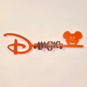May include: Orange-colored decorative key featuring the Disney logo and the word "MAGIC" in a stylized font. The key design includes a Mickey Mouse head at the end, creating a whimsical and recognizable aesthetic.