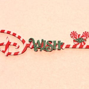 May include: A decorative key-shaped ornament featuring a red and white candy cane striped pattern. The word WISH is written in dark green glittery script in the center, and the bow of the key is a red and white peppermint swirl mouse head.