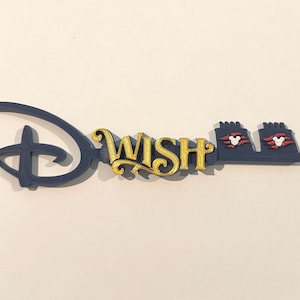 May include: A decorative key-shaped item with a navy blue handle and a gold-colored "WISH" inscription. The key features the Disney logo and Mickey Mouse details, with red and white accents. The item is set against a white background.