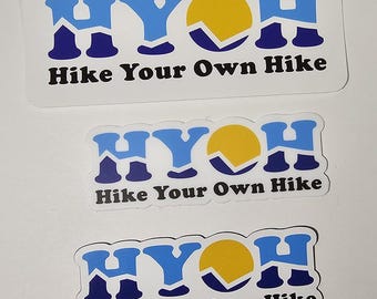 Op de afbeelding: Drie stickers met de tekst "HYOH Hike Your Own Hike". De stickers tonen een gestileerde bergketen met een gele zon op de achtergrond.