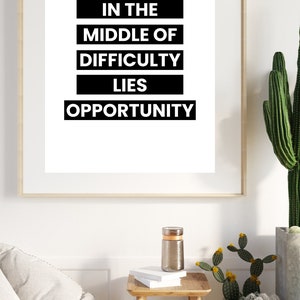 以下が含まれることがあります： 白い背景に「IN THE MIDDLE OF DIFFICULTY LIES OPPORTUNITY」という引用句が太字の黒いテキストで書かれた額入りのプリント。 フレームは明るい木材です。 プリントは白い壁に掛けられており、小さな木製のテーブルの上にあります。