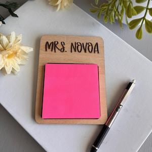 May include: A wooden notepad holder with the text "MRS. NOVOA" engraved on the top. It holds a bright pink sticky note pad. A silver and black pen rests on the side, ready for use. The notepad holder is on a white surface.