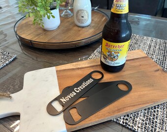 May include: A wooden cutting board with a white marble accent, featuring two black bottle openers with the text "Novoa Creations." A bottle of Sprecher Cream Soda sits next to the openers.