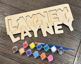 May include: Wooden puzzle with the name "LAYNEY" in capital letters. The puzzle pieces are unpainted. Includes a set of paint pots in various colors and two paintbrushes. The puzzle is designed for painting.