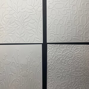 May include: Four white textured paper squares. One features a leaf pattern, another has numbers, a third shows flowers, and the last has a lace-like design. The embossed designs create a tactile and visual appeal.
