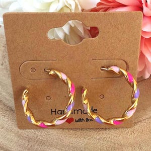 Könnte beinhalten: Goldfarbene Creolen mit gedrehtem Design, mit rosa, lila und hellrosa Emaille-Akzenten. Die Ohrringe werden auf einer braunen Ohrringkarte mit dem Text "Har made with lov" präsentiert.