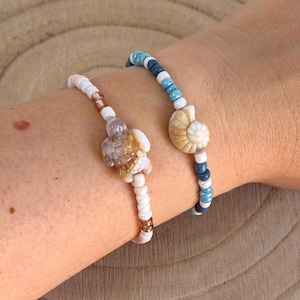 Könnte beinhalten: Zwei Armbänder mit Perlen. Eines hat einen Schildkrötenanhänger, das andere eine Muschel. Die Armbänder haben weiße, braune und blaue Perlen. Der Schildkrötenanhänger ist braun und weiß. Der Muschelanhänger ist beige und weiß.