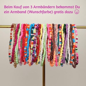 Könnte beinhalten: Eine Sammlung bunter Perlenarmbänder, die an einer goldenen Metallstange hängen. Die Armbänder bestehen aus verschiedenen einfarbigen, durchscheinenden und metallischen Rocailles in Rosa, Blau, Gelb, Grün, Lila, Orange, Rot, Weiß und Braun. Beim Kauf von 3 Armbändern bekommst Du ein Armband (Wunschfarbe) gratis dazu.