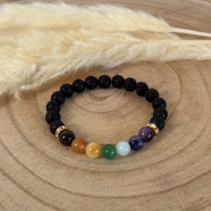 Könnte beinhalten: Schwarzes Lava-Stein-Armband mit einer Reihe farbiger Chakra-Perlen. Das Armband hat rote, orange, gelbe, grüne, hellblaue, dunkelblaue und violette Perlen. Goldfarbene Abstandshalter sind ebenfalls enthalten.