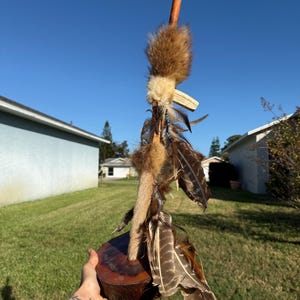 May include: A handcrafted decorative item featuring a wooden base and a central wooden rod adorned with feathers, fur, and other natural elements. The item has a rustic aesthetic with a mix of brown, tan, and cream colors.