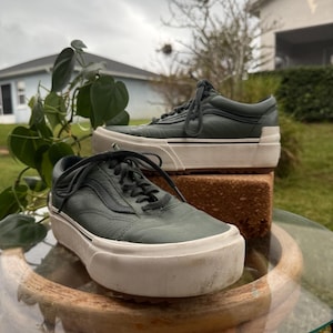 Könnte beinhalten: Dunkelgrüne Sneaker mit weißen Sohlen und Schnürsenkeln. Die Schuhe stehen auf einem Glastisch mit Holzsockel. Im Hintergrund befinden sich ein Ziegelstein und Grün. Die Sneaker haben ein klassisches, niedriges Design.