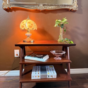 May include: A three-tiered wooden side table with tapered legs. The top surface holds a floral glass lamp and a green plant in a glass vase. The middle shelf holds two books and a pink glass bowl. The bottom shelf holds a large book.