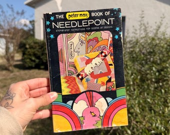 May include: A vintage book titled "The Peter Max Book of Needlepoint." The cover features colorful psychedelic art with the book's title in bold white letters. The book is held in front of a blurred outdoor background.