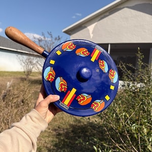 以下が含まれることがあります： 木製のハンドルと蓋が付いた青いエナメル鍋。鍋はカラフルな抽象的なデザインで装飾されています。ハンドルは木製です。「peter max」という文字が鍋の側面に印刷されています。