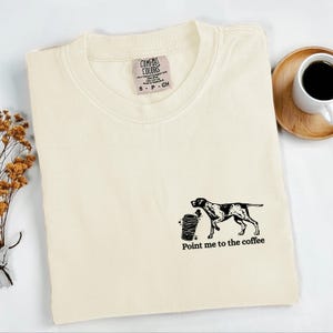 Op de afbeelding: Crèmekleurig Comfort Colors T-shirt met een zwarte afbeelding van een hond en een koffiekopje. De tekst "Point me to the coffee" staat eronder. Een klein label is zichtbaar bij de kraag. Een kopje koffie staat op een houten onderzetter.