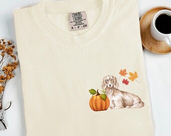 Op de afbeelding: Crèmekleurig T-shirt met een aquarel illustratie van een teckel, een pompoen en herfstbladeren. Het shirt heeft een Comfort Colors label en is netjes opgevouwen. Een kopje koffie staat op een houten onderzetter op de achtergrond.