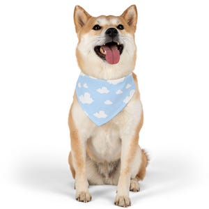 Op de afbeelding: Een Shiba Inu-hond draagt een lichtblauwe bandana met witte wolkenpatronen. De hond zit en kijkt omhoog met zijn mond open en tong uit. De hond heeft bruine en witte vacht.