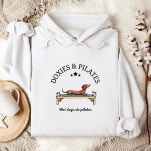 May include: White hoodie with a drawstring hood. The front features a graphic of a dachshund on a Pilates reformer, with the text "DOXIES & PILATES" above and "Hot dogs do pilates" below. The design is in black and brown.