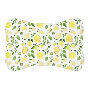 Puede incluir: Cama para mascotas en forma de hueso con fondo blanco y un patrón repetido de limones amarillos, hojas verdes y flores blancas. Los limones están enteros, en rodajas y en racimos. La cama tiene bordes redondeados.