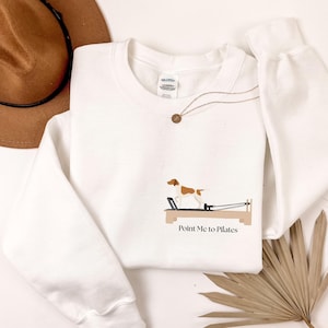 Könnte beinhalten: Weißes Sweatshirt mit einer Grafik eines Hundes auf einem Pilates-Reformer-Gerät. Der Text "Point Me to Pilates" befindet sich unter der Grafik. Ein brauner Hut und eine goldene Halskette sind ebenfalls zu sehen.