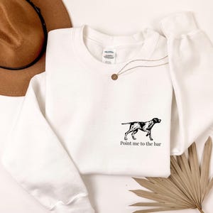 Op de afbeelding: Witte sweatshirt met een zwart-witte hondenafbeelding en de tekst "Point me to the bar". Een gouden ketting en een bruine hoed zijn zichtbaar. De sweatshirt is gemaakt van zacht materiaal.