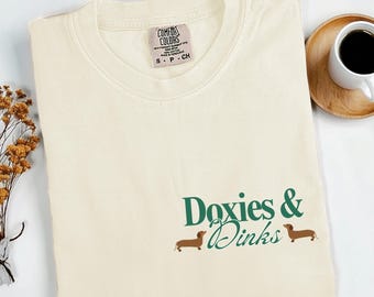 May include: Cream-colored t-shirt with the words "Doxies & Dinks" in green script and two brown dachshund illustrations. The shirt has a Comfort Colors tag and is folded neatly. A cup of coffee sits on a wooden coaster.