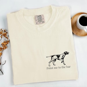 May include: Cream-coloured t-shirt featuring a black and white dog graphic and the text "Point me to the bar." The shirt is folded and has a Comfort Colors tag. A cup of coffee sits on a wooden coaster.