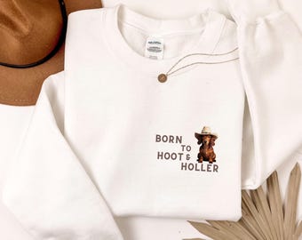 May include: White sweatshirt with a brown dachshund wearing a cowboy hat and the text "Born to Hoot & Holler".