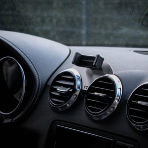 Peut inclure: Tableau de bord de voiture avec une surface noire semblable à du cuir et des détails argentés. Le tableau de bord comprend des bouches d'aération avec des garnitures chromées, un support de téléphone et un compteur de vitesse. Gros plan.