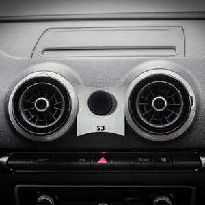 Può includere: Primo piano dell'interno di un'auto con due bocchette di ventilazione rotonde e un pulsante nero con la scritta "S3" su di esso. Le bocchette di ventilazione sono argentate e hanno lame nere. Il pulsante si trova al centro del cruscotto.