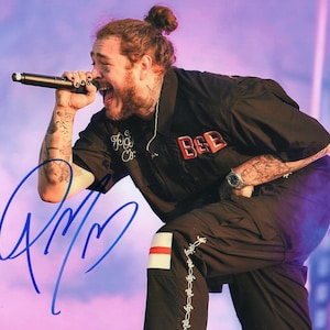 May include: A black and white photo of a man with a beard and a bun in his hair. He is wearing a black shirt with the word "BEEB" on it and black pants. He is holding a microphone in his right hand and singing. The photo is signed with a blue signature.