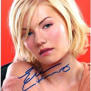 May include: A close-up portrait of a woman with blonde hair, wearing a light brown shirt. She is looking at the camera with a slight smile. Her signature is written in blue ink on the shirt.