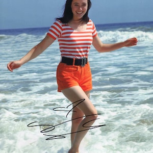 May include: A woman in a red and white striped shirt and orange shorts stands in the ocean waves. She is smiling and has her arms outstretched. The photo is signed by the woman.