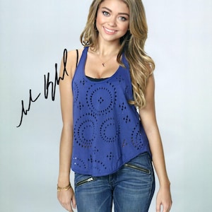 May include: A young woman with long blonde hair is wearing a blue tank top with a geometric pattern and blue jeans. She is smiling and looking at the camera. The tank top has a scoop neck and a racerback style. The jeans have a zipper detail on the front pockets.