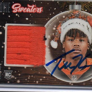 May include: A trading card featuring Tee Higgins of the Cincinnati Bengals. The card has a swatch of orange fabric and a photo of Higgins wearing a Santa hat. The card is labeled "Rookie Sweaters."