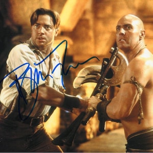 May include: A signed photograph featuring two men from the movie "The Mummy." One man is holding a weapon, while the other is looking on. The image has a warm color palette, with the actors in costume.