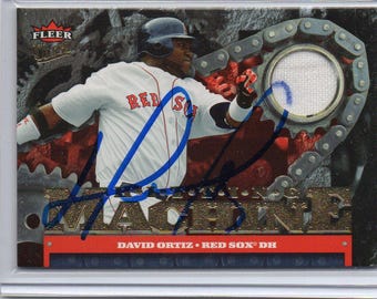 May include: A baseball trading card featuring David Ortiz of the Boston Red Sox. The card has a metallic background with a gear-like design and a patch. The player is in a white jersey with "RED SOX" on it. The card is signed in blue ink.