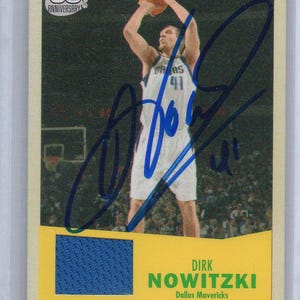 May include: A Topps 50th Anniversary basketball card featuring Dirk Nowitzki of the Dallas Mavericks. The card shows Nowitzki in action, shooting a basketball. The card has a yellow border and a blue jersey swatch.