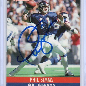 May include: A Pro Set NFL card featuring Phil Simms, quarterback for the Giants, in a blue and white uniform throwing a football. The card has the NFL logo and the text "The Official NFL Card". The card is signed.